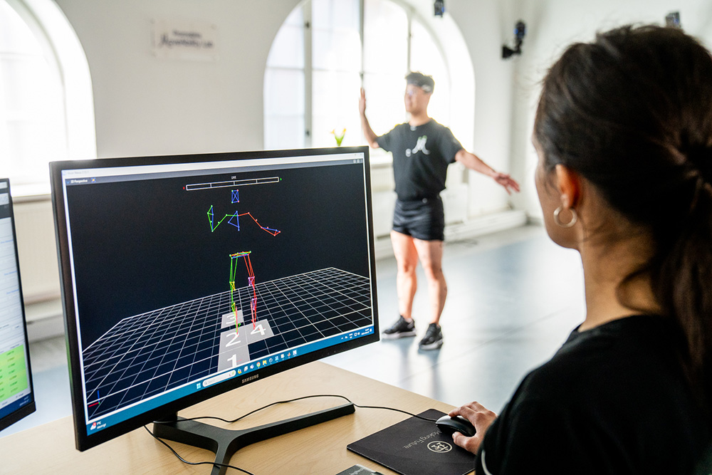 Person vid skärm, i bakgrunden stående man i kortbyxor.
