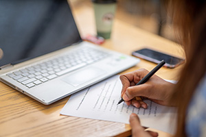 A hand writing on a piece of paper in front of a computer.